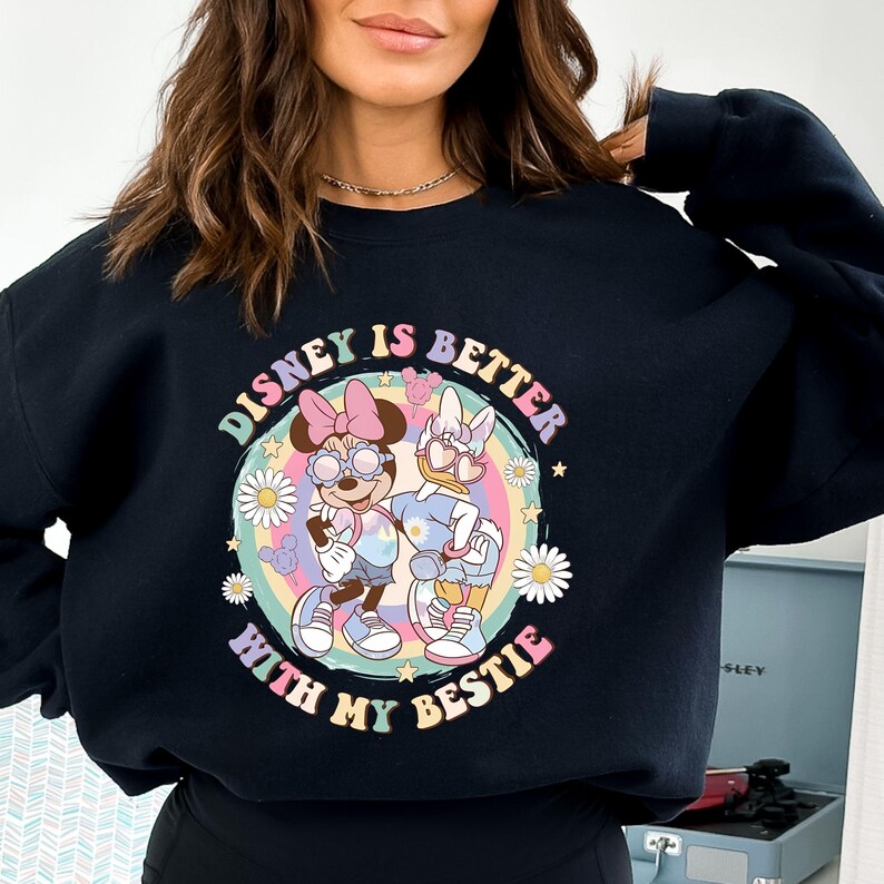 May include: Black sweatshirt featuring a colorful graphic of Minnie Mouse and Daisy Duck with the text "DISNEY IS BETTER WITH MY BESTIE". The design includes a rainbow, daisies, and stars, creating a cheerful and playful aesthetic.