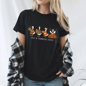 Puede incluir: Camiseta negra con un gr&aacute;fico de cuatro gansos con atuendos oto&ntilde;ales. Los gansos llevan comida y rastrillan hojas, con el texto "JUST A THANKFUL GOOSE" debajo. La camiseta est&aacute; hecha de un material suave.