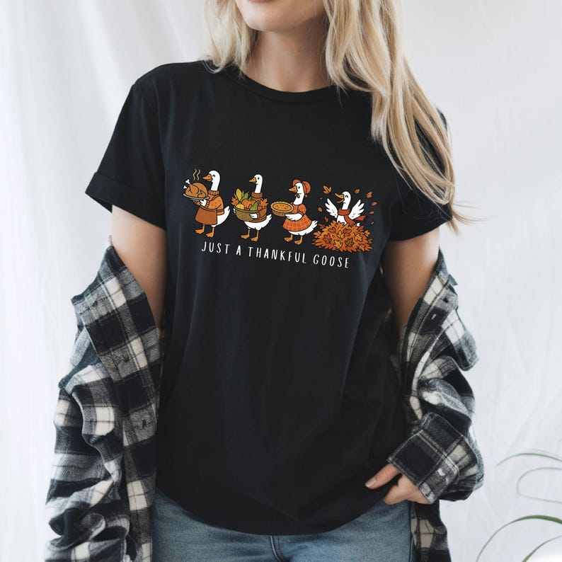 Puede incluir: Camiseta negra con un gr&aacute;fico de cuatro gansos con atuendos oto&ntilde;ales. Los gansos llevan comida y rastrillan hojas, con el texto "JUST A THANKFUL GOOSE" debajo. La camiseta est&aacute; hecha de un material suave.