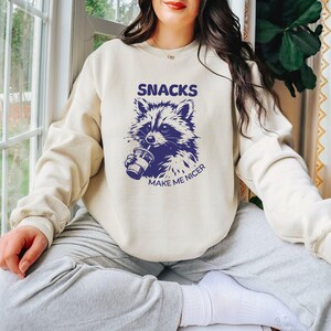 May include: A cream-coloured sweatshirt featuring a navy blue graphic of a raccoon drinking from a cup. The text "SNACKS" is above the raccoon, and "MAKE ME NICER" is below. A casual, comfortable style.