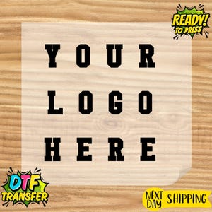 May include: A rectangular DTF transfer sheet featuring the black text YOUR LOGO HERE in a bold, blocky varsity font. The design is centered on a semi-transparent film. In the top right corner, a green and yellow graphic reads READY TO PRESS.