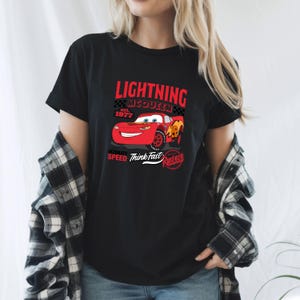 May include: Black t-shirt featuring a graphic of Lightning McQueen from the movie Cars. The design includes the text "Lightning McQueen est. 1977" and "Ultimate Speed Think Fast". The car is red with yellow and orange accents.