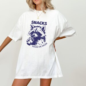 May include: White t-shirt featuring a navy blue raccoon graphic drinking from a cup. The text "SNACKS" is above the raccoon, with "MAKE ME NICER" below. The shirt has short sleeves and a loose fit.