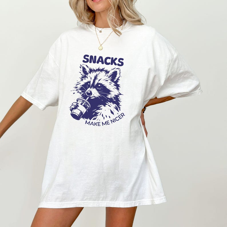 May include: White t-shirt featuring a navy blue raccoon graphic drinking from a cup. The text "SNACKS" is above the raccoon, with "MAKE ME NICER" below. The shirt has short sleeves and a loose fit.