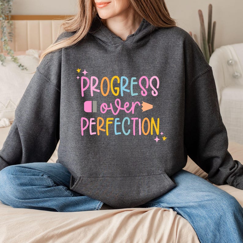 May include: Dark gray hoodie with the text "PROGRESS over PERFECTION" in colorful, playful lettering. The design includes a pink eraser, an orange pencil, and yellow stars. The hoodie has a front pocket and a hood.