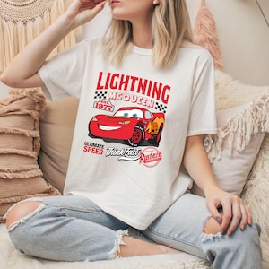 May include: White t-shirt featuring a graphic of Lightning McQueen from the movie Cars. The design includes the text "LIGHTNING McQUEEN est. 1977" and the phrases "ULTIMATE SPEED", "Think Fast", and "Rusteze". The cartoon car is red with the number 95.