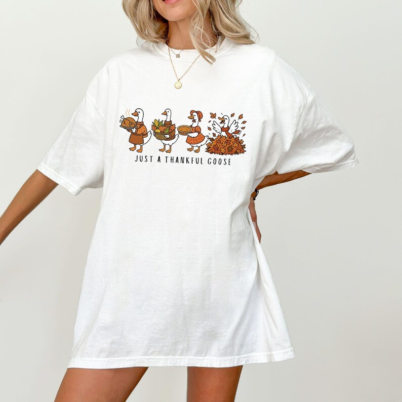 Puede incluir: Camiseta blanca con un gr&aacute;fico de tres gansos llevando elementos de Acci&oacute;n de Gracias. Los gansos visten trajes marrones y naranjas, con el texto "JUST A THANKFUL GOOSE" debajo.