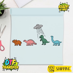 May include: A light blue DTF transfer sheet featuring four cartoon dinosaurs in brown, pink, green, and blue, with a UFO above the green dinosaur. The sheet has the text "READY! TO PRESS" and "DTF TRANSFER".