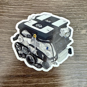 May include: A sticker featuring a detailed illustration of a car engine. The engine is primarily black and white with accents of yellow and blue. The Nissan logo is visible on the top. The sticker is on a wooden surface.