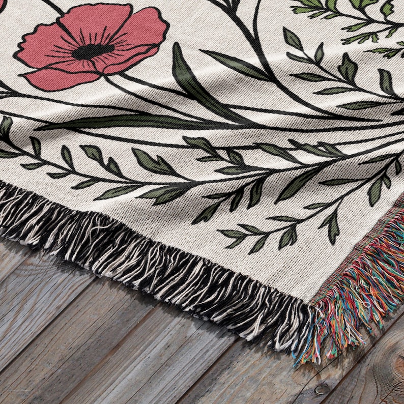 May include: Cream-colored woven throw blanket with a floral design featuring a large pink poppy and green leaves. The blanket has a black fringe along the edge and a multicolored fringe on the corner. The blanket is on a wooden surface.