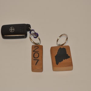 May include: Wooden keychains with silver rings. One keychain has the number "207" engraved in black. The other keychain features a black outline of the state of Maine. A black car key is attached to the keychains.