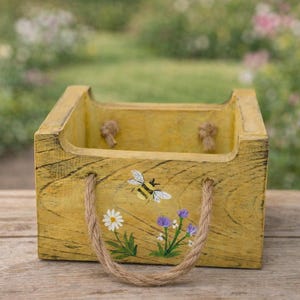 May include: Yellow wooden box with a rope handle and a painted bee and flower design. The box has a rustic, distressed finish and is suitable for storage or decoration. The background is a blurred view of greenery.