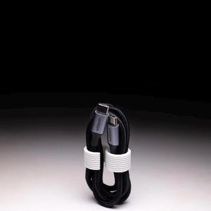 May include: A black USB-C charging cable, coiled and secured with two white bands. The cable has gray connectors with the brand name "UGREEN" printed on them. The background is a gradient of black and white.