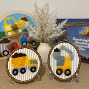 May include: Two circular wooden embroidery hoops featuring needle-felted construction vehicles on white and gray striped fabric. One hoop displays a yellow dump truck with a brown load, and the other shows a yellow excavator with a gray arm.