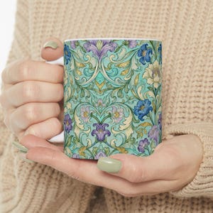 May include: A white ceramic mug featuring a detailed floral pattern in shades of teal, purple, blue, and cream. The design includes stylized flowers and foliage, creating a vintage aesthetic. The mug is held in a person's hands.