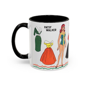 May include: A white ceramic mug with a black interior and handle, featuring a vintage Patsy Walker paper doll illustration. The design includes a red-haired figure, a green dress, a yellow and orange skirt, and the text Patsy Walker in black.