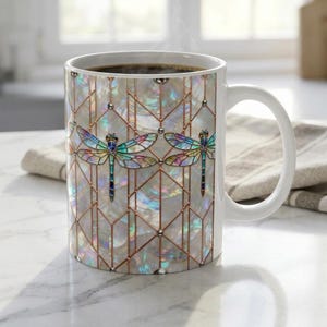 May include: White ceramic coffee mug featuring a decorative design of two dragonflies with iridescent wings. The dragonflies are set against a geometric pattern with copper-colored lines and a pearlescent background. The mug has a white handle.