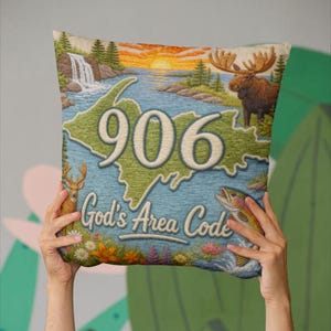 May include: Decorative pillow featuring a map of the 906 area code with a scenic landscape. The pillow includes the text "God's Area Code", a moose, a deer, and a fish. The design is in shades of green, blue, and beige.