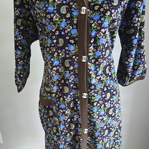 May include: A vintage floral print robe with a dark blue background, adorned with blue, green, and tan floral patterns. The robe features a brown trim, three-quarter sleeves, and is fastened with silver buttons.