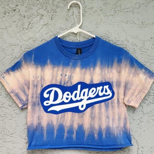 May include: A blue tie-dye crop top with the word "Dodgers" in white script across the front. The shirt has a crew neck and short sleeves. The tie-dye effect features a light pink and blue pattern.