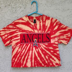 May include: Red and tan tie-dye crop top with the word "ANGELS" in a white rectangle, and a red "A" with a halo. The shirt is hanging on a blue hanger against a textured gray background.