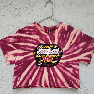 May include: A short-sleeved crop top featuring a burgundy and cream tie-dye spiral pattern. The center displays a black graphic patch with the text Commanders in white and a yellow W logo, surrounded by decorative yellow, white, and burgundy lines.
