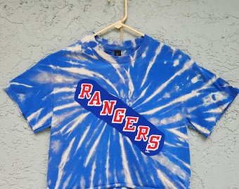 Puede incluir: Una camiseta corta de color azul y blanco con estampado tie-dye que presenta una banda diagonal azul en el pecho con la palabra RANGERS impresa en letras rojas con bordes blancos. La prenda se muestra en una percha de plástico beige.