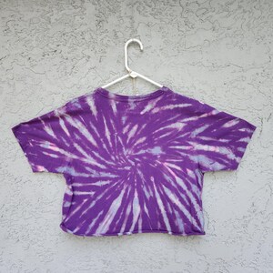 May include: A purple tie-dye crop top with a spiral design. The shirt is hanging on a white hanger against a textured gray background. The tie-dye pattern features shades of purple, white, and light pink.