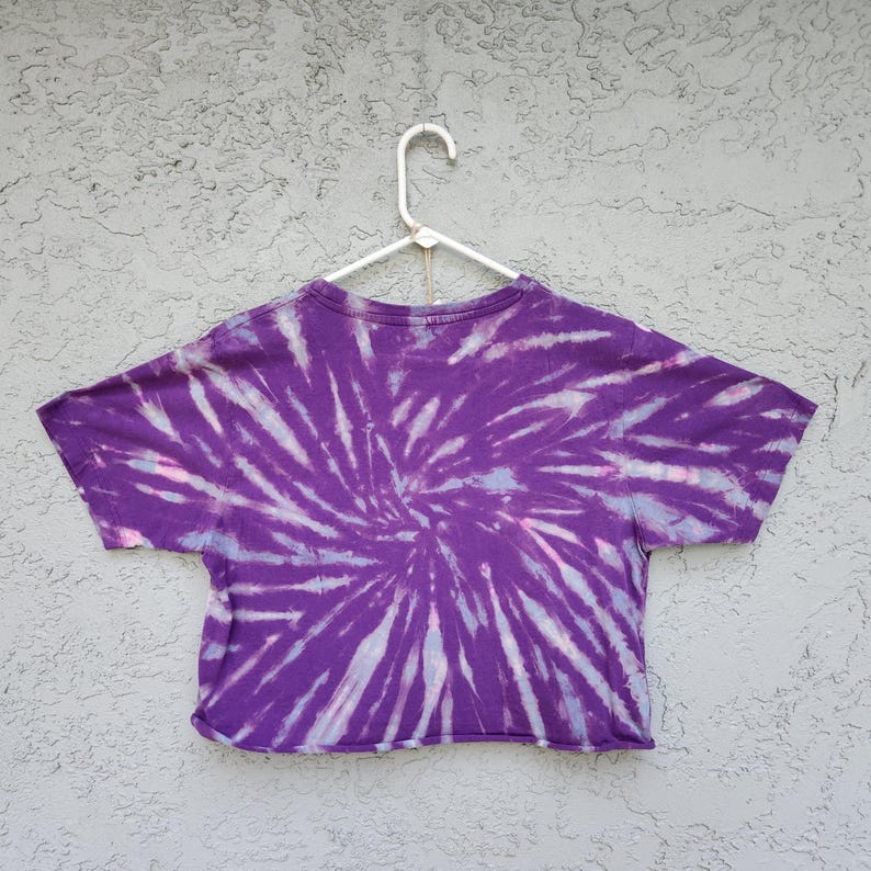 May include: A purple tie-dye crop top with a spiral design. The shirt is hanging on a white hanger against a textured gray background. The tie-dye pattern features shades of purple, white, and light pink.