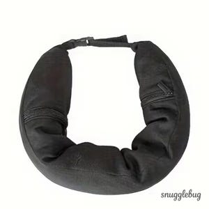 May include: Black travel neck pillow with a zip pocket and adjustable buckle fastening. The pillow is designed to wrap around the neck for support and comfort. The brand name "snugglebug" is visible in the bottom right corner.