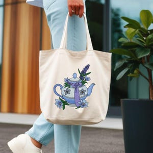 May include: A cream-colored canvas tote bag featuring an embroidered design of a light blue teapot decorated with purple flowers and a purple ribbon. The teapot is surrounded by sprigs of lavender and additional blue floral accents.