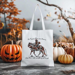 Könnte beinhalten: Weiße Tragetasche mit einem Cowboy- und Pferdedesign, verziert mit floralen Mustern. Die Tasche trägt den Text: "Don't just follow your dreams, chase them with everything you've got." Kürbisse und Herbstlaub sind im Hintergrund.