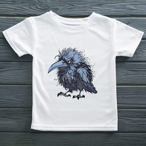 Puede incluir: Una camiseta blanca de manga corta con una ilustración gráfica central de un cuervo azul oscuro y gris, de aspecto despeinado, con un pico afilado y una expresión intensa. El ave tiene un plumaje detallado y desordenado, y se apoya sobre dos garras.