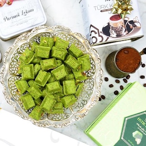 May include: Overhead shot of a silver platter filled with green pistachio sweets. Several tins of Turkish delight and a box of pistachio sweets are visible. A small copper pot of coffee and coffee beans are also in the image.
