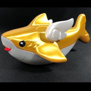 May include: A gold shark ornament with white wings, a white underside, and a red tongue. The shark has a black eye and is set against a dark background. This decorative item is a unique piece of art.
