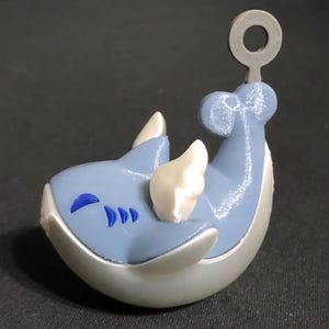 May include: A light blue and white figurine of a sea creature with a loop at the top. The figurine has a light blue body with white accents and a silver base. The creature has a smiling face and three blue markings.