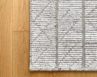 Puede incluir: Primer plano de una alfombra rectangular con una superficie texturizada blanquecina y un patrón de cuadrícula gris geométrico. La alfombra está colocada sobre un suelo de madera clara.