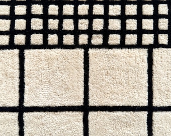 May include: A tufted rug featuring a geometric grid pattern with a high-pile texture. The design consists of large, cream-coloured squares separated by thick, black lines, transitioning into a section of smaller, dense cream squares.