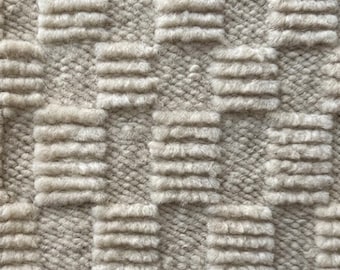 May include: Close-up of a textured, off-white rug. The rug features a repeating pattern of raised, rectangular blocks, creating a three-dimensional effect. The blocks are arranged in a grid-like formation, adding visual interest to the neutral-toned textile.