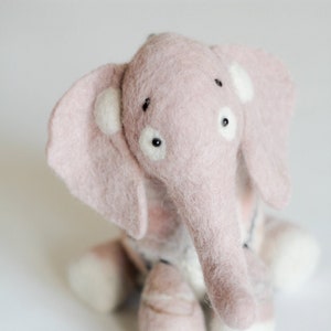 May include: A handmade pink felt elephant toy with white accents on its ears and feet. The elephant has a long trunk, small black eyes, and is sitting on a white surface. The toy is soft and plush.