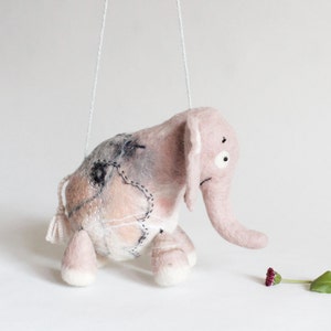 May include: A handmade pink felt elephant puppet with white string attached. The elephant has a long trunk, large ears, and stitched details. The puppet is suspended in the air, with a small flower and leaf on the ground.
