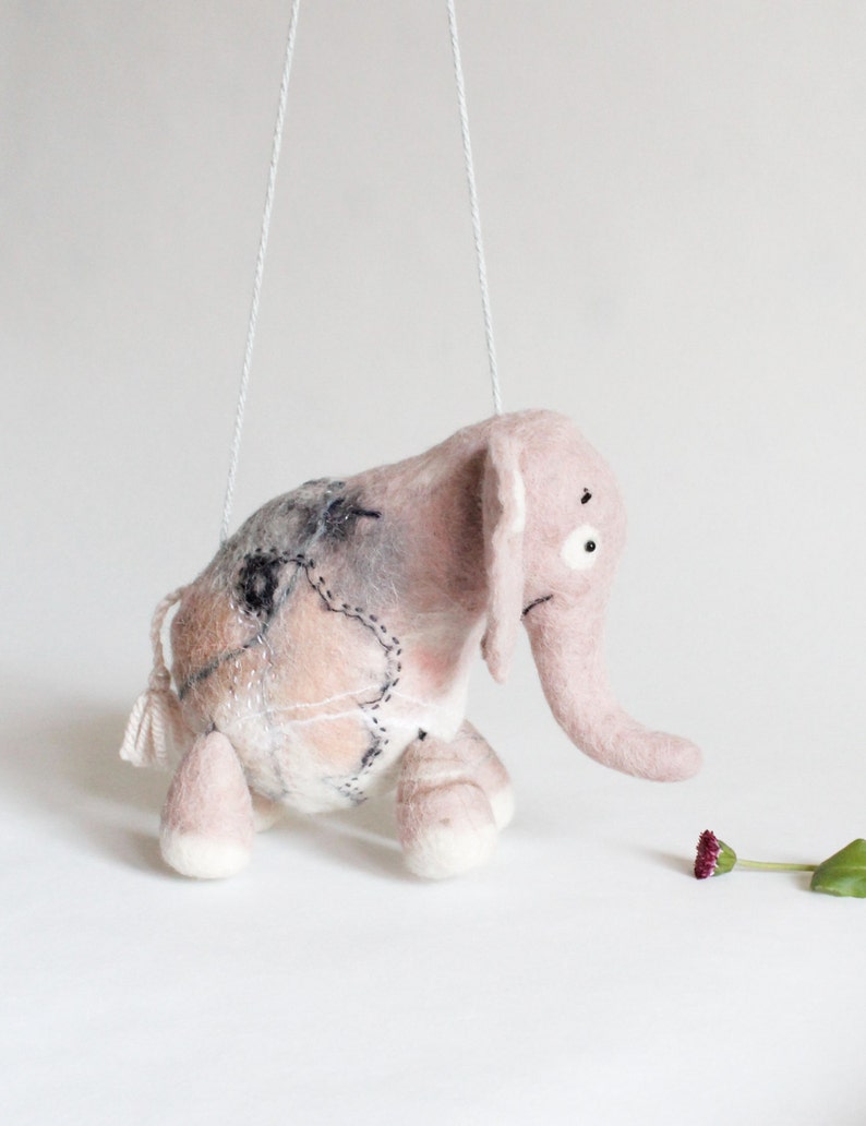 May include: A handmade pink felt elephant puppet with white string attached. The elephant has a long trunk, large ears, and stitched details. The puppet is suspended in the air, with a small flower and leaf on the ground.