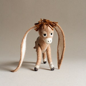 Puede incluir: Figura de burro hecha a mano en fieltro en tonos marrón y beige. El burro tiene orejas largas y caídas con interior blanco, un hocico blanco y ojos oscuros. La melena es de un marrón más oscuro y se apoya sobre cuatro patas con pezuñas negras.