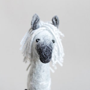 Könnte beinhalten: Handgefertigte Pferdfigur aus Filz. Das Pferd hat einen grauen Körper, eine schwarze Schnauze und schwarze Hufe. Seine Mähne und sein Schopf bestehen aus weißem Garn. Die Ohren des Pferdes sind grau, und es hat kleine schwarze Augen. Das Spielzeug ist eine weiche Skulptur.