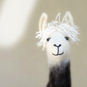 May include: A handcrafted llama figurine with a white head and shaggy fringe. The llama's neck transitions from white to dark brown. It has small black eyes and a stitched smile, creating a whimsical appearance. The background is a soft, neutral color.