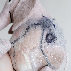 May include: A handmade pink and gray felt elephant toy with stitched details. The elephant has large ears, a trunk, and a small tail. The body features decorative stitching in various colors, including black, white, and pink.