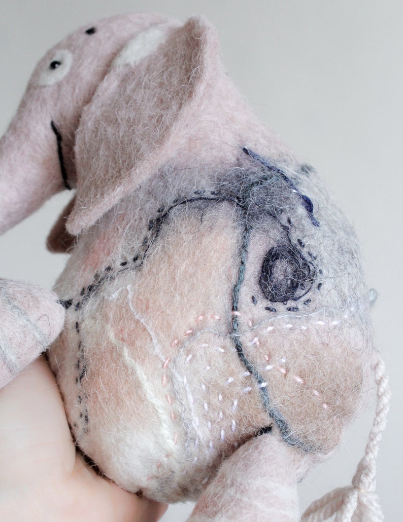 May include: A handmade pink and gray felt elephant toy with stitched details. The elephant has large ears, a trunk, and a small tail. The body features decorative stitching in various colors, including black, white, and pink.