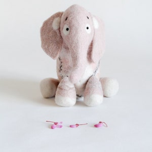 May include: A handmade pink felt elephant toy with white accents on its ears and feet. The elephant has black eyes and is sitting on a white surface. Small pink flower petals are scattered in front of the elephant.