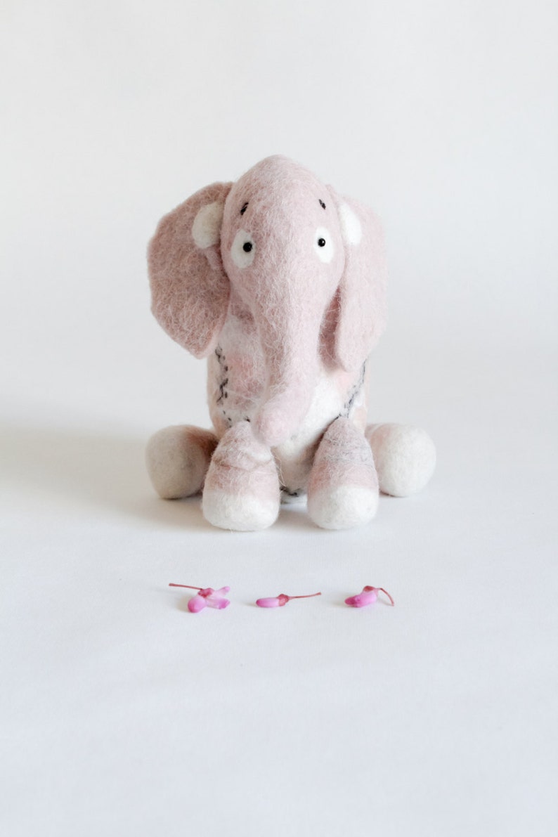 May include: A handmade pink felt elephant toy with white accents on its ears and feet. The elephant has black eyes and is sitting on a white surface. Small pink flower petals are scattered in front of the elephant.