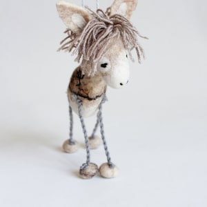 May include: A handmade felt donkey ornament with a light brown body, long ears, and a fringe of brown yarn for a mane. The donkey has gray string legs with small ball feet. The ornament hangs from a white string.